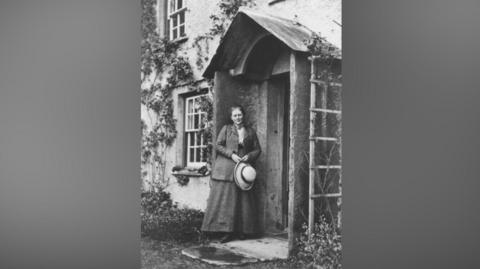 A black and white image of Beatrix Potter at her Hill top home in 1913. She is standing in the porch of a cottage-style home. Her hair is tied back and she is holding a hat with a wide brim in her hands. She is wearing a coat and skirt. The cottage has sash windows and there are creepers going up the walls and shrubs surrounding the house. 