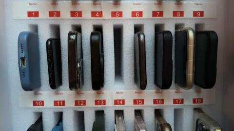 Rows of mobile-phones sat in small phone-shaped holes within a locking case. There are black, blue, and gold phone cases visible in-between the white polystyrene holes.