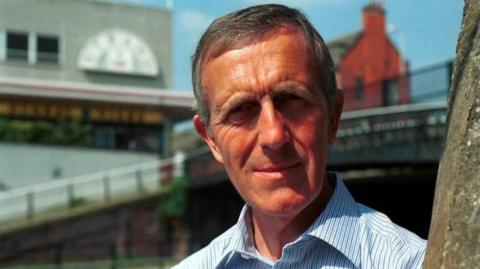 Albert Kirby is looking at the camera. He has short brown/grey hair and is wearing a striped blue shirt. The blurred exterior of the shopping centre is in the background.