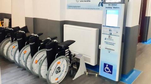 A row of folded wheelchairs in a docking station at King's College Hospital with a rental station to the side. 