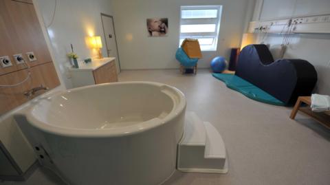 A birthing pool suite at another NHS hospital - which includes a white tub placed inside a room with a chair and a table, inflatable ball, mat and a white window. There is a picture of a baby on the wall.
