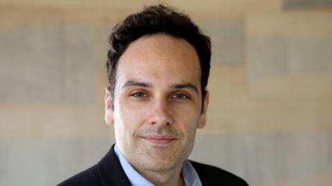 Dr Javier Andreu-Perez is smiling. He has black hair and stubble. He is wearing a black suit jacket and a pale blue shirt. There is a wall behind him, although it has been blurred.