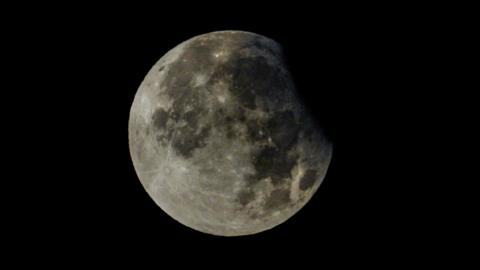 The Moon is seen with a tiny part of the top-right covered by a partial eclipse. The Moon looks white in a clear sky.