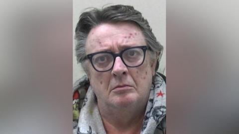 Mugshot of Storey. He is clean shaven with thick long grey hair swept back and various red spots and marks on his face. He is wearing black glasses and a grey hoody with colourful characters on it, possibly the Grinch.