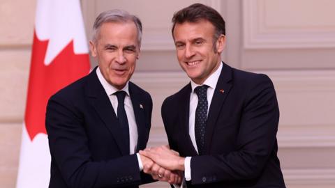 Mark Carney and Emmanuel Macron shake hands while smiling and looking at the camera in front of a Canadian flag during a press conference in Paris on Monday. They both wear a suit and tie.