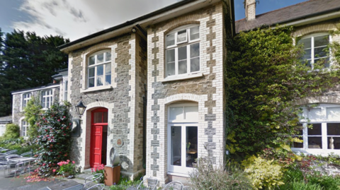 A picture of the outside of the hotel. The building is all brick, grey and white, and has a red door. There is plants surrounding the hotel with hedges next to it.