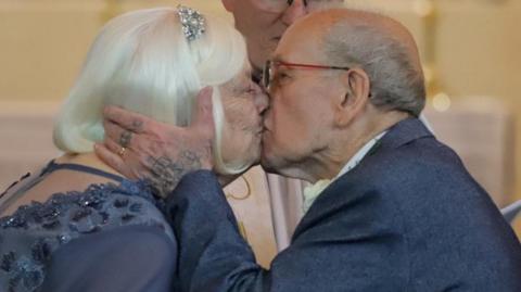 Kath and Terry kiss in church after getting married, with the vicar looking on