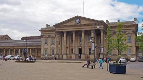 A photo of the outside of Huddersfield station. There is a large pedestrianised area in front of the Victorian building. A number of people are milling around and there are a few taxis parked at either side of the building.