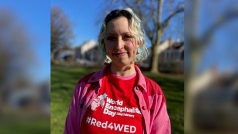 Shareen Elnagy wearing a red World Encaphalitis Day t-shirt and a pink and red cardigan over the top. She is smiling at the camera and has her blonde hair up, with trees and grass behind her.