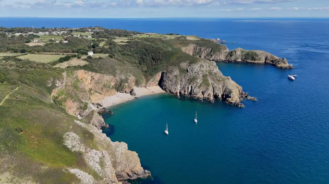 An aerial image of the island of Sark, showing a beach and cliff faces under sunny skies. There re a number of boats bobbing in the sea.
