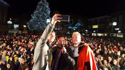 Joe Clarke takes a selfie while standing next to a man holding a microphone and another man in mayoral robes. Behind them is a large crowd