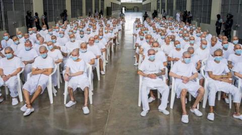 A group of hundreds of men with shaved heads, wearing white t-shirts and shorts or trousers and blue surgical masks, are seen sitting on white plastic chairs in an open space at a confinement centre in El Salvador