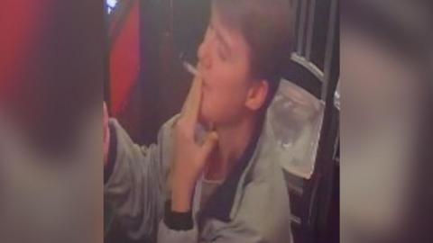 CCTV image of a young man smoking a cigarette and wearing a grey bomber jacket looking up at the wall of a bar