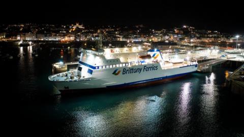 A large Brittany Ferries vessel docked at a brightly lit Guernsey harbour at night. Surrounding the ship are illuminated port facilities and buildings with St Peter Port town visible in the background under a dark sky. 