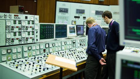 Two men with their backs to the camera looking at a control panel with lots of button and switches.