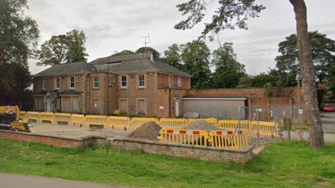 Google Maps view of Manor House in Royal Wootton Bassett. It is a red brick building. A yellow construction fence and construction vehicles and machinery can be seen outside the building.