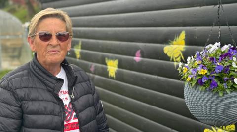 Hamilton resident Sandra Panton - a blonde woman in a black jacket, standing next to a black wooden shed with a hanging basket of purple flowers