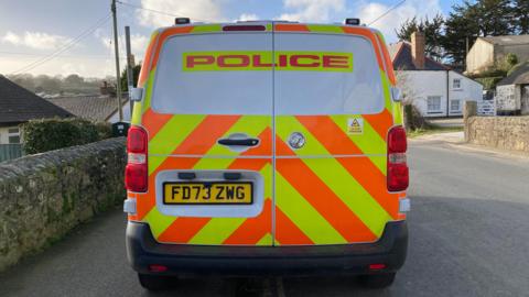 A stock image of a police van pictured from behind. The van is parked in a rural village.