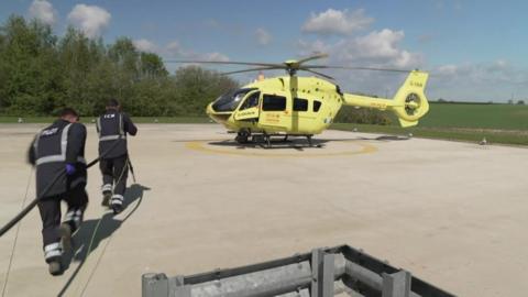Two men wearing dark overalls run a fuel line across to a fluorescent yellow air ambulance. 