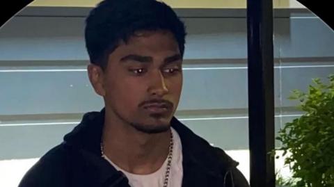 Young man, with dark hair and trimmed moustache poses looking away from camera. He wears a white t-shirt, chain necklace and dark hoodie. He stands in front of a window and a small green plant is positioned to the right of him.