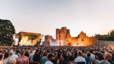 A large crowd is gathered in front of an outdoor concert stage, beside illuminated castle ruins at dusk.