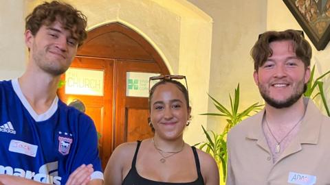 Three young people stand in a row, inside a church with sofas in the background. Jack has a blue Ipswich football jersey on, Freya wears a black strap top and Adam wears a beige polo shirt with a flopping brown middle-parted fringe.