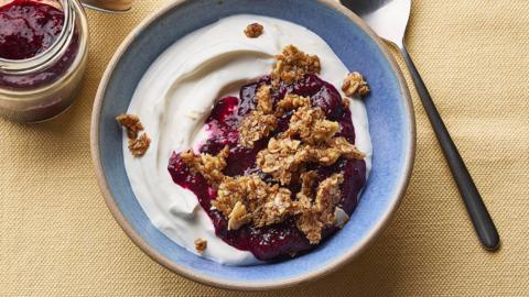 Greek yoghurt in a blue bowl topped with golden granola pieces and red jam