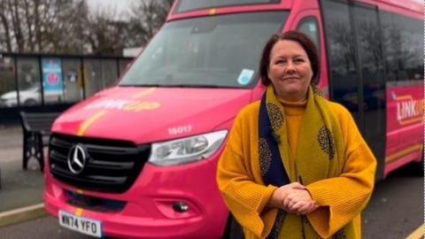 Janice Silvester-Hall is stood in front of a pink mini bus which has white and yellow lettering which reads 'link up' on the bonnet and side.
She is holding her hands together and is wearing a big yellow jumper with a mustard and blue scarf.