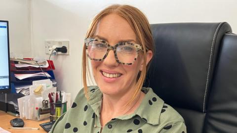 Stacey Lock wearing large-framed leopard-skin glasses and a green shirt with black spots. She is sitting in a black office chair in front of a computer.