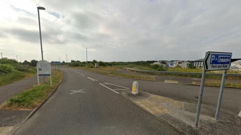 Road with signs on the left verge and on a central island - buildings can be seen on the right.
