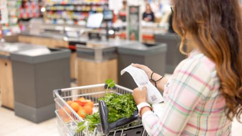 A woman is shown in a grocery store with a trolley that has fruit and vegetables in it. She has her back to the camera, and is looking at a receipt.