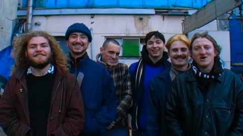 Six male band members are pictured outside what looks like a derelict school building smiling in a row.