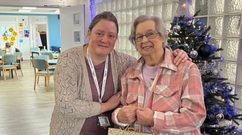 A volunteer from the Age UK Clockhouse Cafe and Jill Rowley. The volunteer has her arm around Jill who is wearing a pink and white shirt. Jill is also holding a paper bag. The pair are standing in front of a Christmas tree.