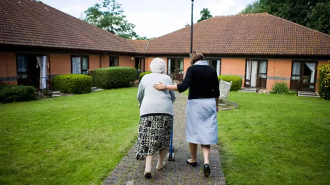 An elderly woman using a walking frame walks with a nurse towards a property 