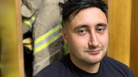 Oliver Dent sat in a fire station locker room in front of high visibility clothing. He has black spiky hair and is wearing a navy T-shirt.