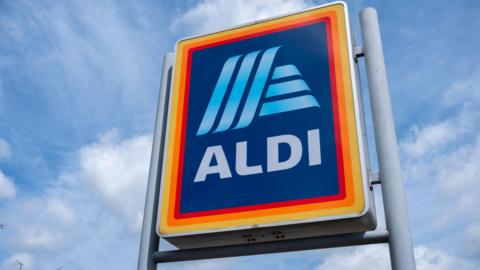 A large sign for Aldi on grey poles set against a blue sky.