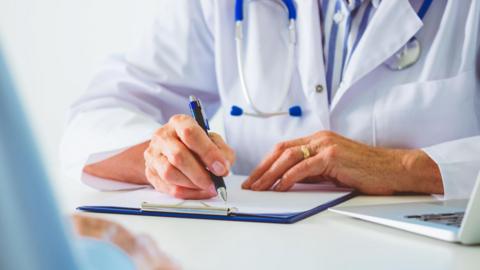 A stock image of a medical professional in a white doctor's coat writing on a clip board