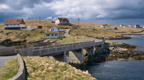 The island communities of Housay and Bruray in the island group of Out Skerries. Houses are scattered across a grassed area with a bridge at the centre of the photograph connecting the two islands and the sea on the right.