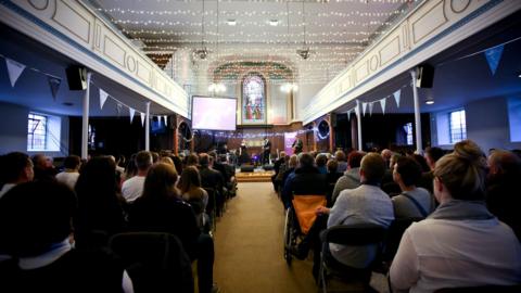 A crowd of seated people watching three musicians perform on stage. There are two balconies either side of the photo leading to the front, with fairy lights strung between them