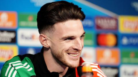 Declan Rice smiles during a press conference - he has short brown hair and wears a sports jacket