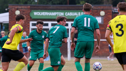 Three footballers in green shirts and two in yellow shirts are on a pitch with little green grass at this angle. A building in the background includes the words Wolverhampton Casuals on a sign.