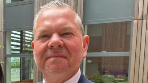 A man with grey hair is wearing a white shirt and blue jacket. A building is behind him.