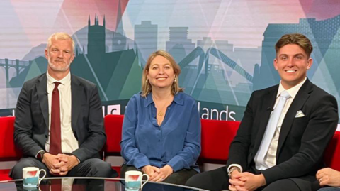 A man with white hair and a beard in a grey suit and red tie next to a woman with blonde hair and a blue blouse and a man with short brown hair and a dark suit with a pale blue tie, all sat on a red sofa