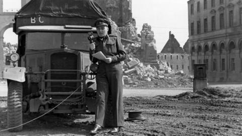 Frank Gillards broadcasts in front of a BBC car. There are ruined buildings in the background