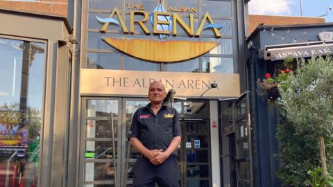 A man standing outside the Alban arena wearing navy shirt and trousers