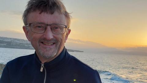 Trevor Stevens is slightly to the left of the picture, smiling and looking into the camera. He is a man in his 60's with dark grey hair. He is wearing rectangular glasses and a navy zip up jacket. There are some rocks in the background, and the sea. Above it, the sun is setting on the horizon. 