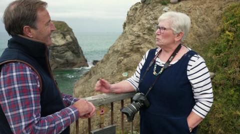 David Braine is wearing a red ad blue checked shirt and dark waistcoat and is standing by a guard rail besides the sea with Julie Taylor. She has a camera with a large lens around her neck. They are talking and a large piece of rock, which is part of Bedruthan Steps is behind them.
