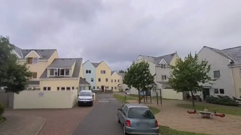 Multiple houses, some painted yellow and one painted blue. There is a grey car parked facing away from the camera and a white van parked facing towards the camera. There is a tree to the left and two trees to the right. 