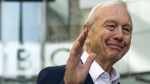 John Humphrys waving goodbye outside BBC's New Broadcasting House. He has white hair and is wearing a white shirt with a navy blazer. 