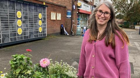 A woman stands in front of a row of shops next to a flowerbed full of plants and flowers.